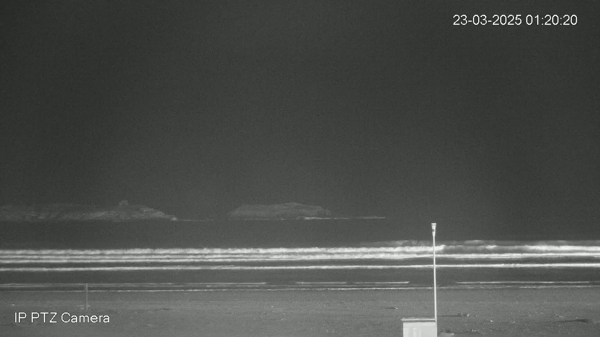 A dark, nighttime beach scene shows calm waves lapping the shore under a clear sky.