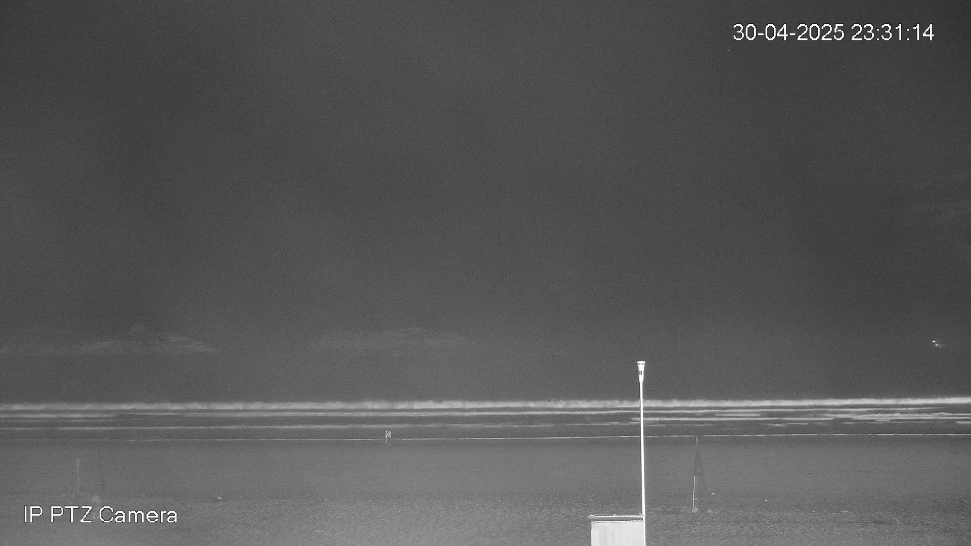 A dark, nighttime beach scene shows a calm ocean with small waves, a few people walking along the shore, and a single lamp post.
