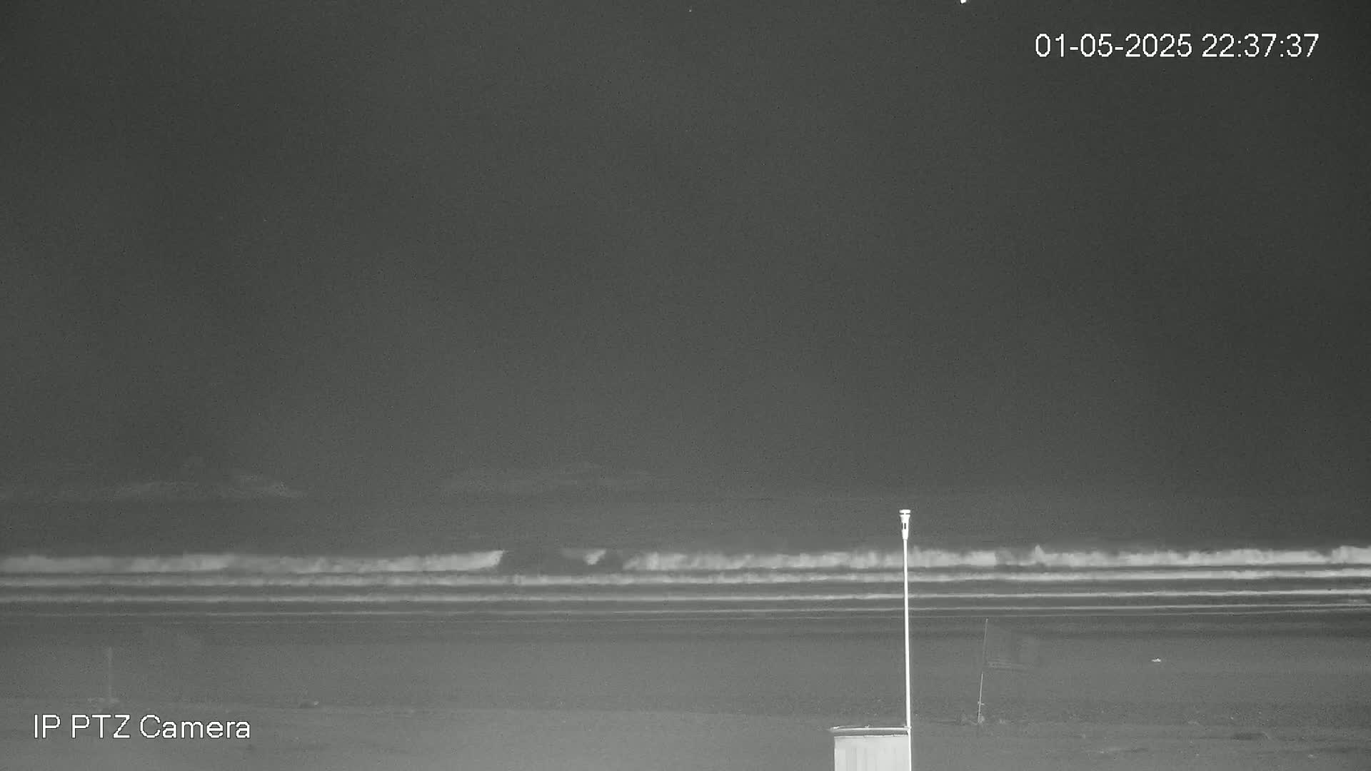 Under a dark night sky, waves crash on a beach near a single pole and small structure.