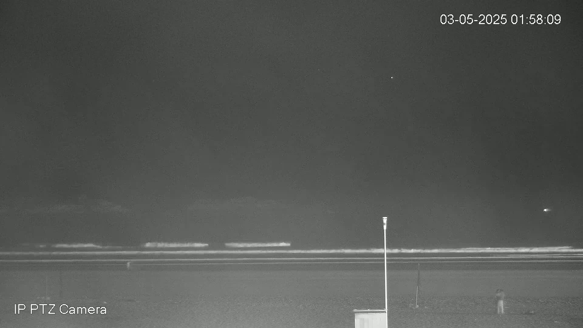 A dark, nighttime beach scene shows gentle waves lapping the shore under a mostly clear sky, with a single person visible in the distance.