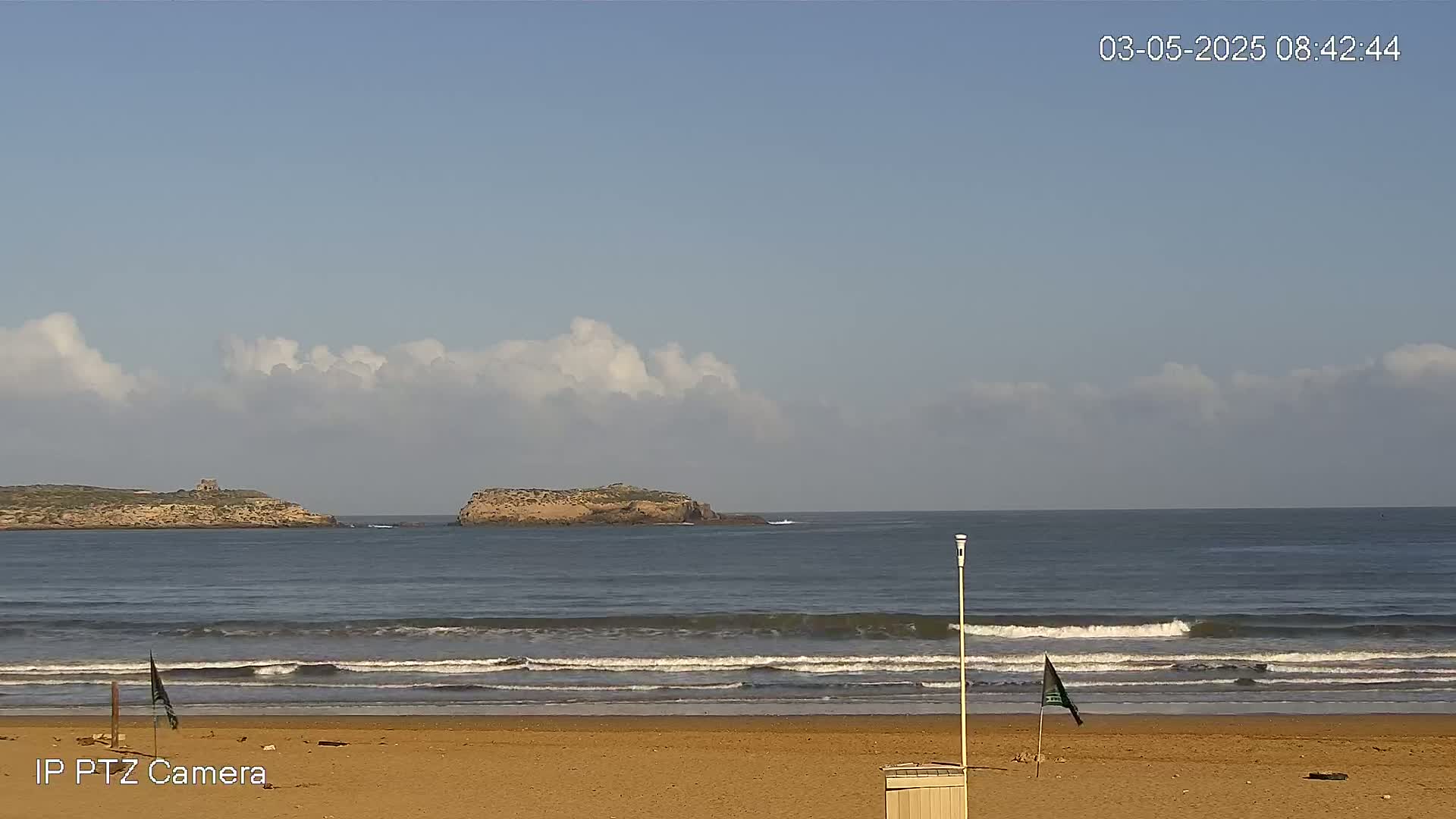 A sandy beach with gentle waves rolls onto the shore under a partly cloudy sky, with small islands visible in the distance.