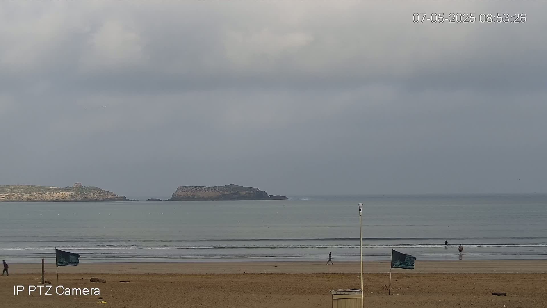 A sandy beach with a few people walking on it stretches out to a calm sea under a cloudy sky, with small islands visible in the distance.