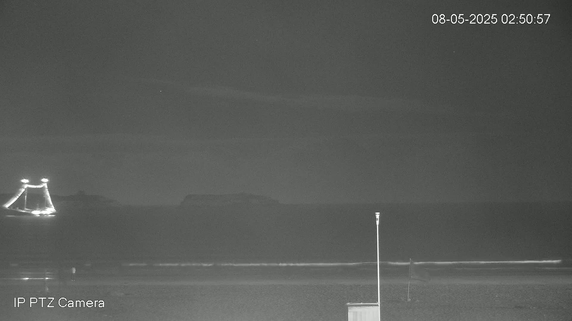 A nighttime beach scene shows a dark ocean with a faint, light-colored shore, a distant structure with lights, and a single pole standing near a small structure on the sand.