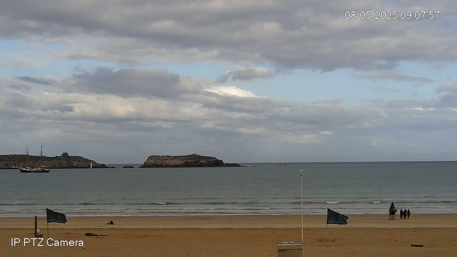 A mostly cloudy sky overlooks a calm ocean with small waves lapping a sandy beach where a few people and a camel are visible near the waterline and a sailboat is seen further out at sea.