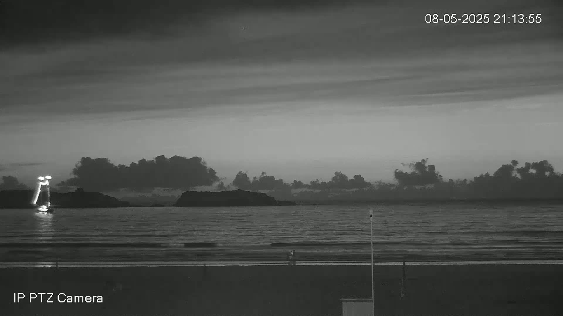 A grayscale image shows a dark, cloudy evening over a calm ocean with a beach in the foreground and a distant island or headland.