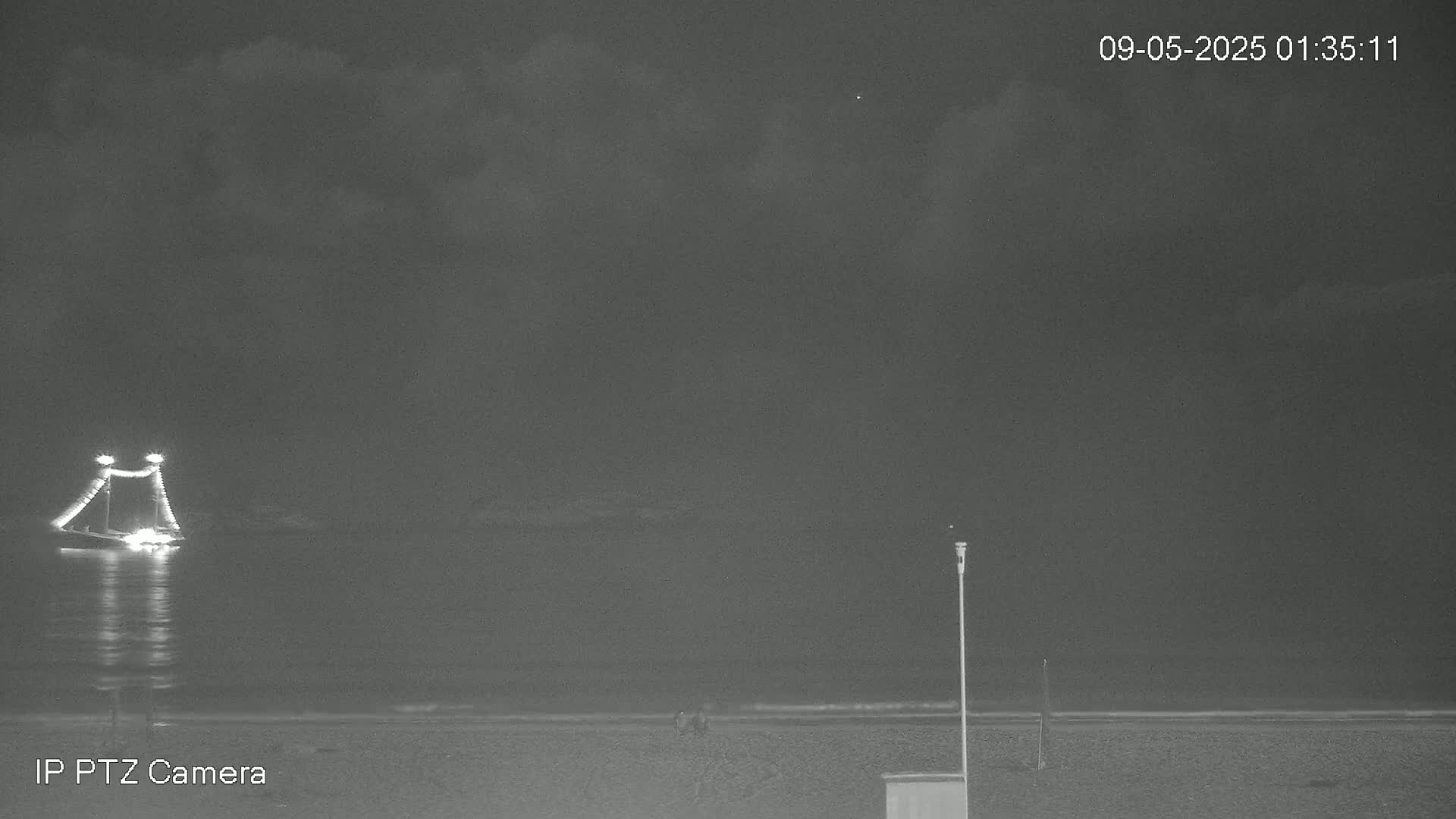 A nighttime, cloudy scene shows a lit-up boat on the water near a dark beach with a few people visible in the distance.