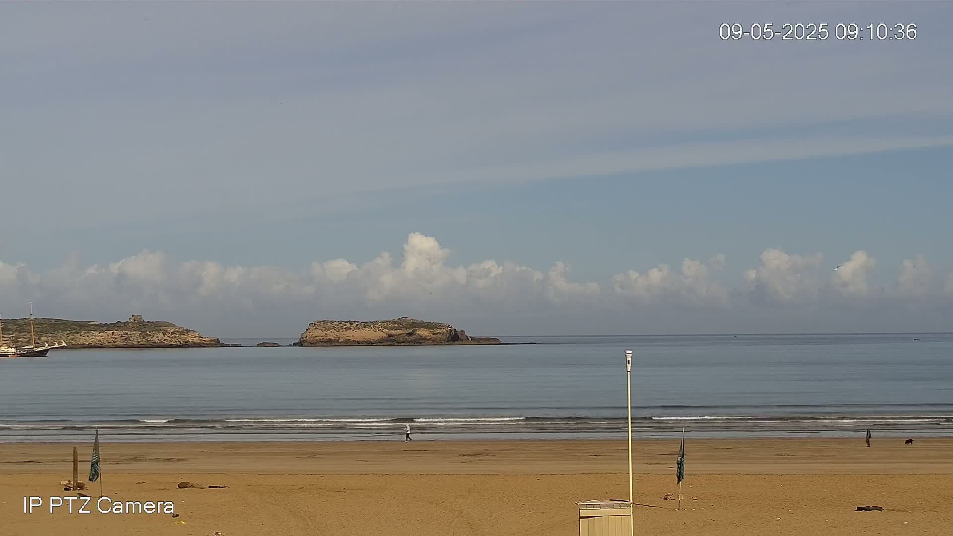 A sandy beach with gentle waves laps the shore under a mostly cloudy sky, with islands visible in the distance.
