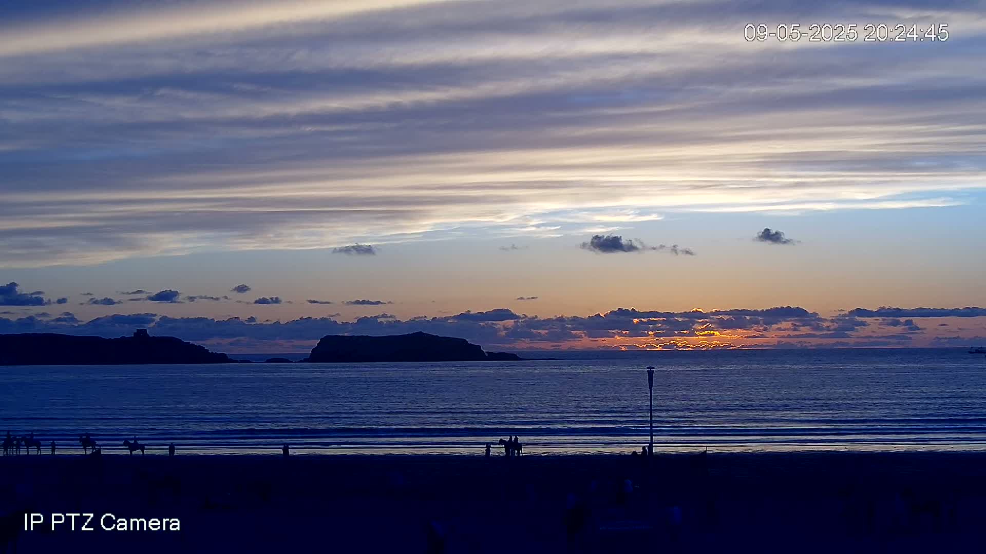 A sunset over a calm ocean, with a beach in the foreground and silhouetted islands in the distance under a partly cloudy sky.