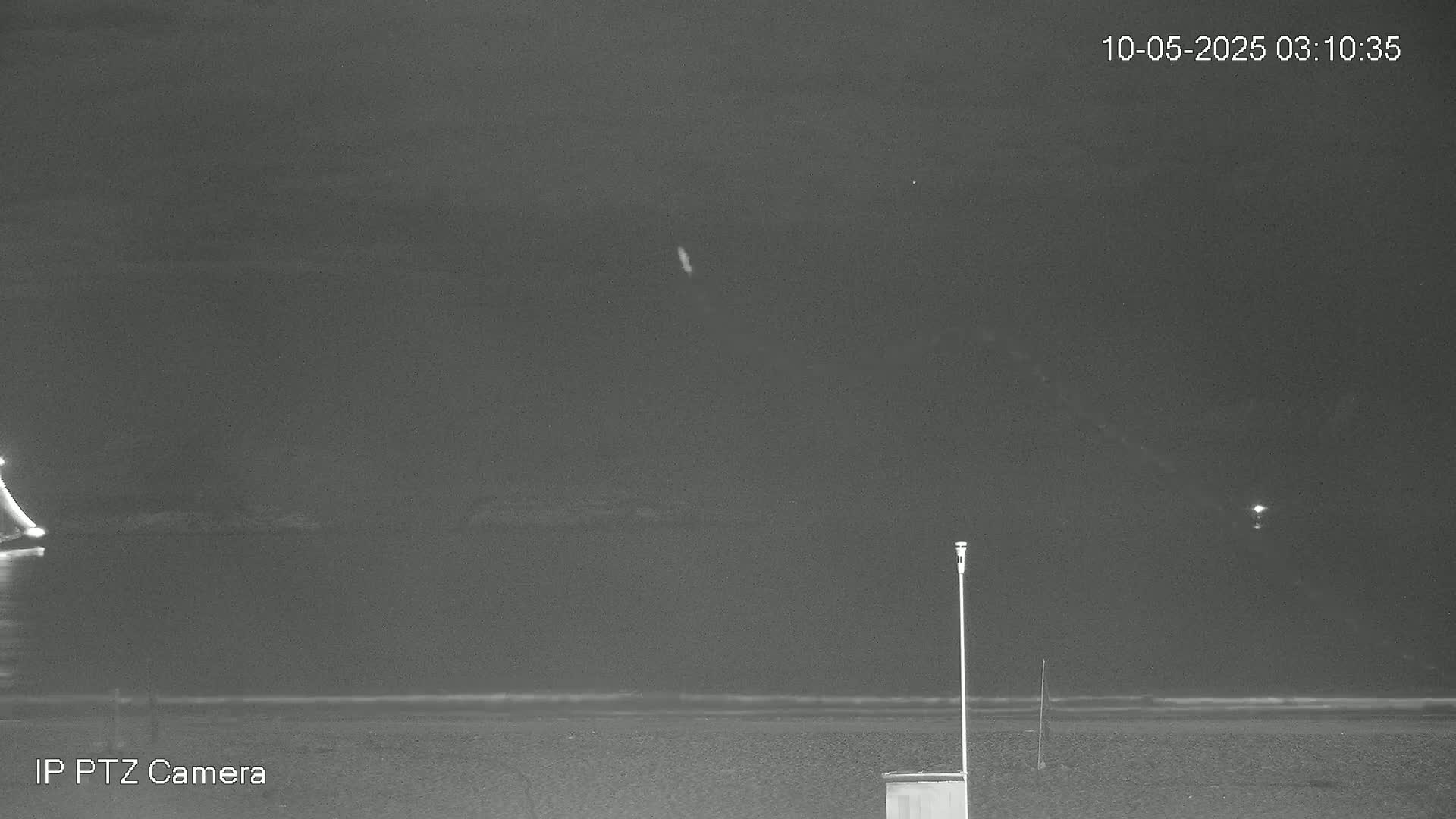 A nighttime, low-light image shows a dark body of water with a calm surface, a sandy beach, and a tall, thin pole in the foreground.