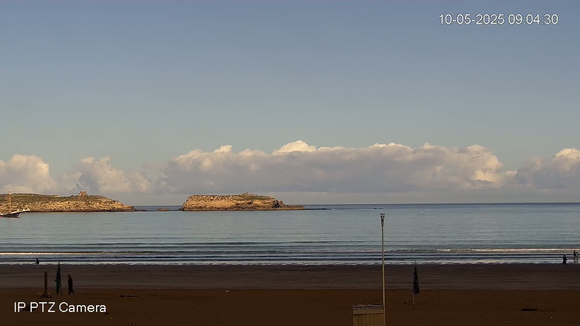 A sandy beach with a few people on it stretches to a calm ocean with small waves, under a partly cloudy sky, and two small islands visible in the distance.
