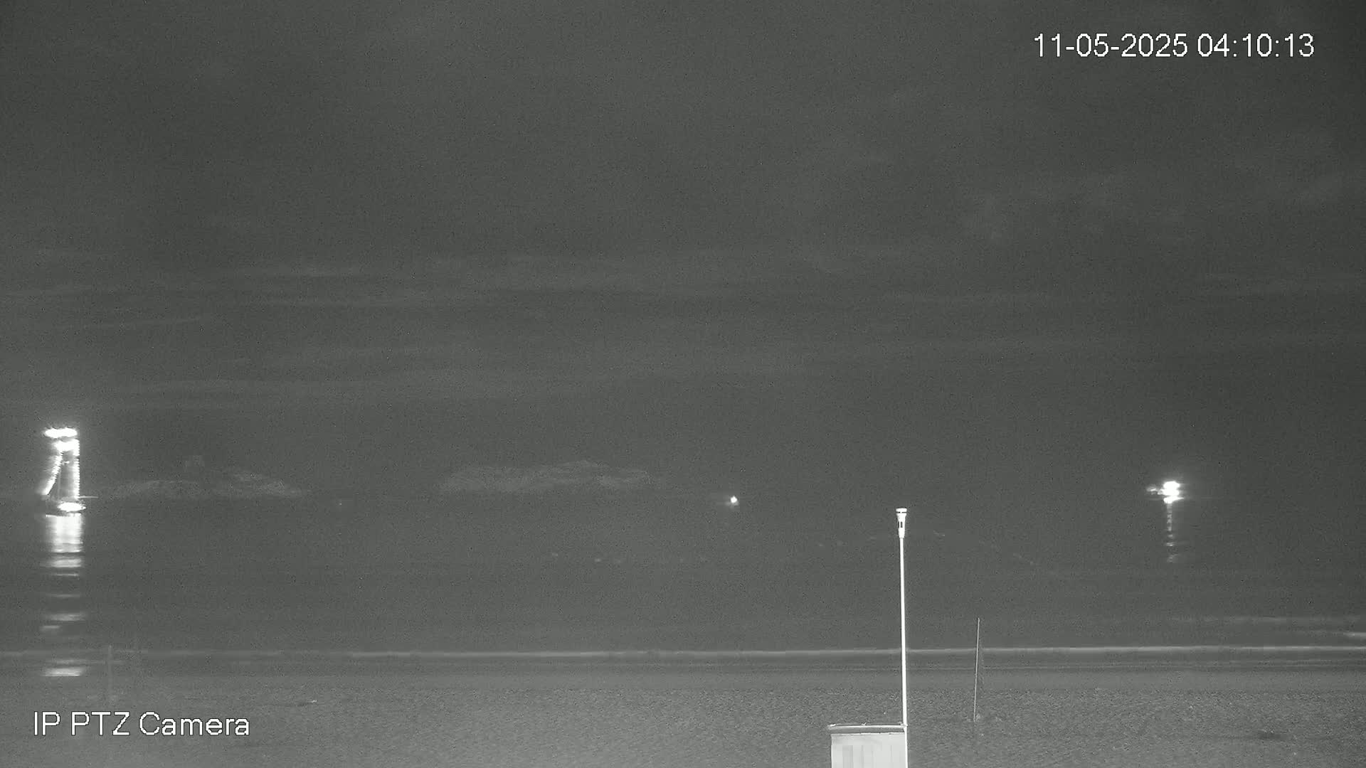 A nighttime, dark grayscale view of a beach with a calm ocean, lit by distant lights.