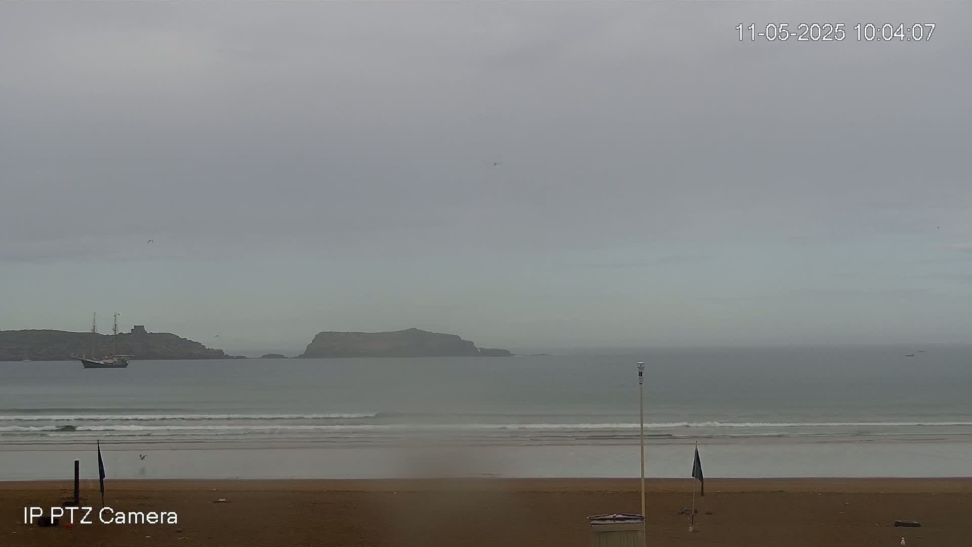 A hazy, overcast day reveals a sandy beach with small waves lapping the shore, a sailboat at sea, and a distant island.