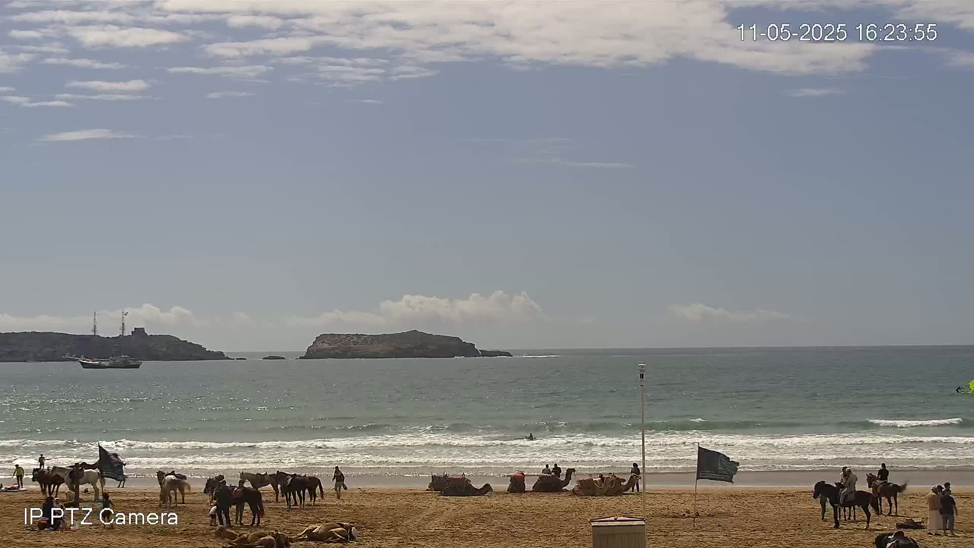 A sandy beach with people and horses is visible, with a calm ocean and islands in the distance under a partly cloudy sky.