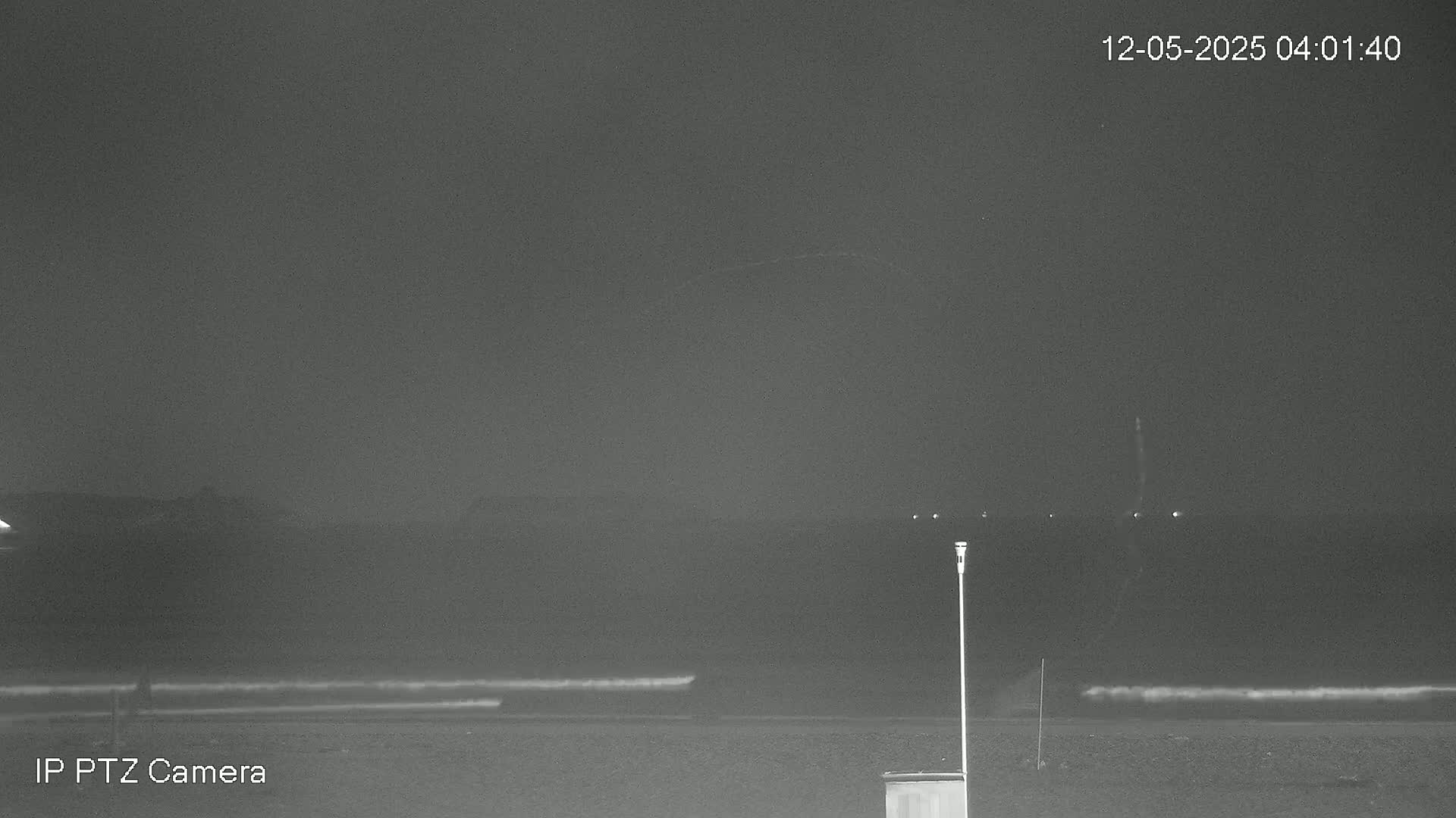 A nighttime beach scene shows dark water with small waves, a dark shoreline, and a dark sky with a faint, thin cloud.