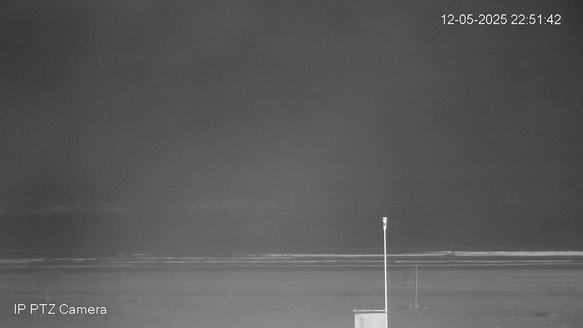 A dark, nighttime beach scene shows a calm ocean with a single light post and a small structure on the sand under an overcast sky.