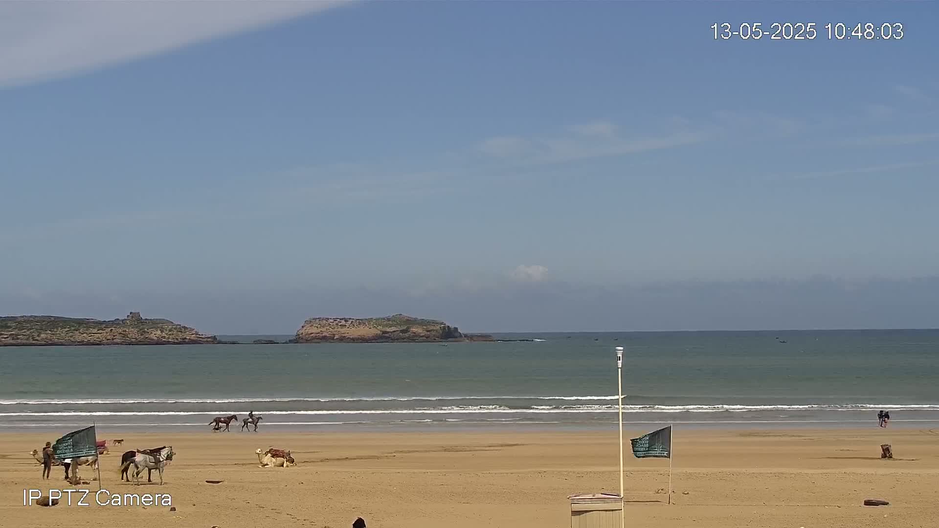 A sandy beach with several people riding horses and camels, under a mostly clear blue sky, overlooks a calm ocean with two small islands in the distance.