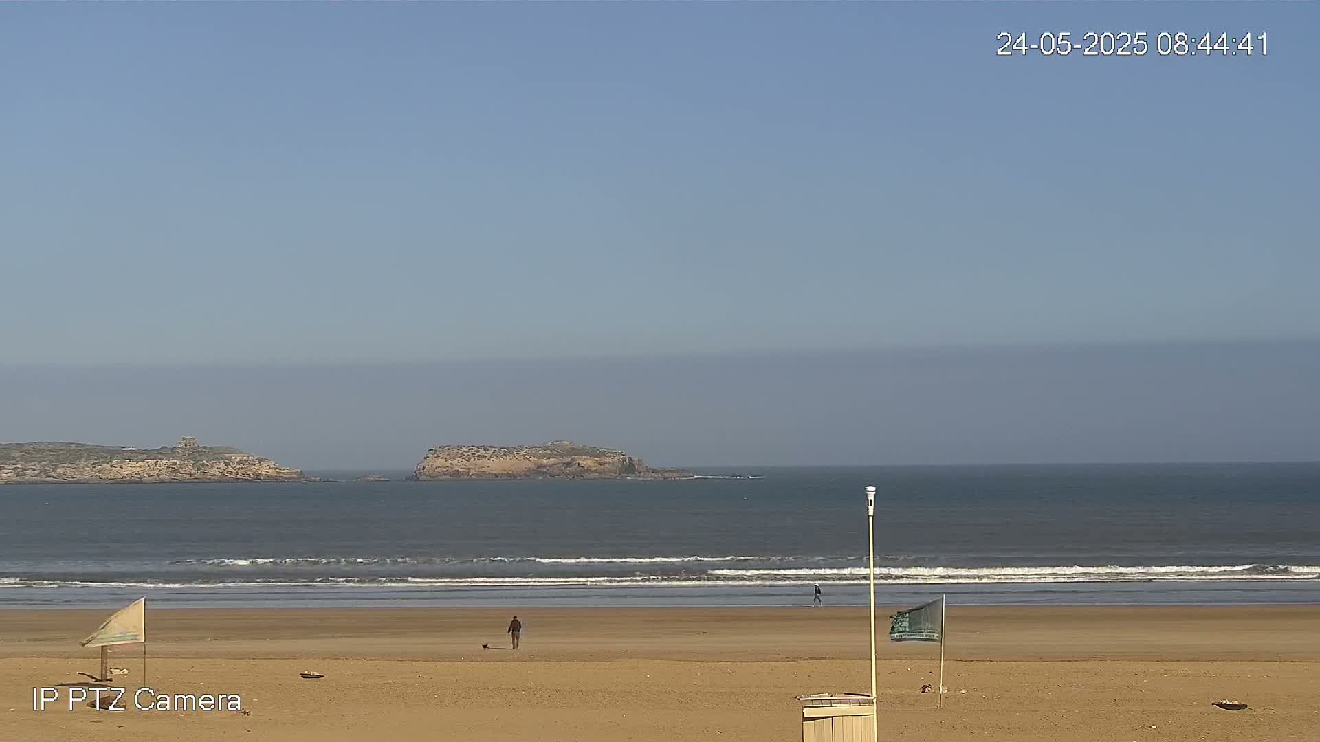 A sandy beach with a few people walking, small waves lapping the shore, and two small islands visible in the distance under a clear, light blue sky.