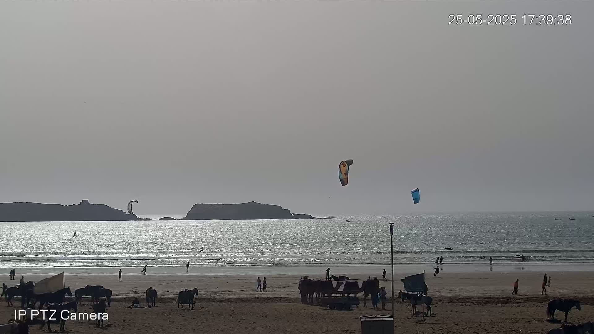 A hazy beach scene shows people on horseback and people swimming and kitesurfing in the ocean, with a rocky island visible in the distance.