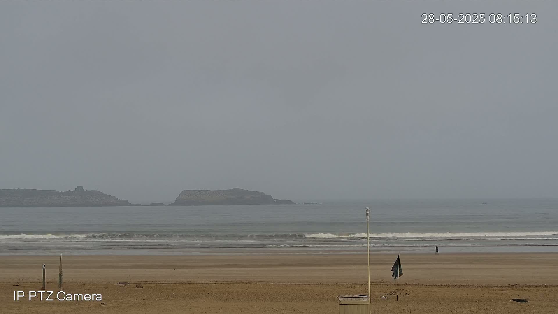 A hazy, overcast day shows a wide sandy beach with gentle waves lapping the shore, and a lone person walking near the waterline with distant rocky outcrops visible on the horizon.
