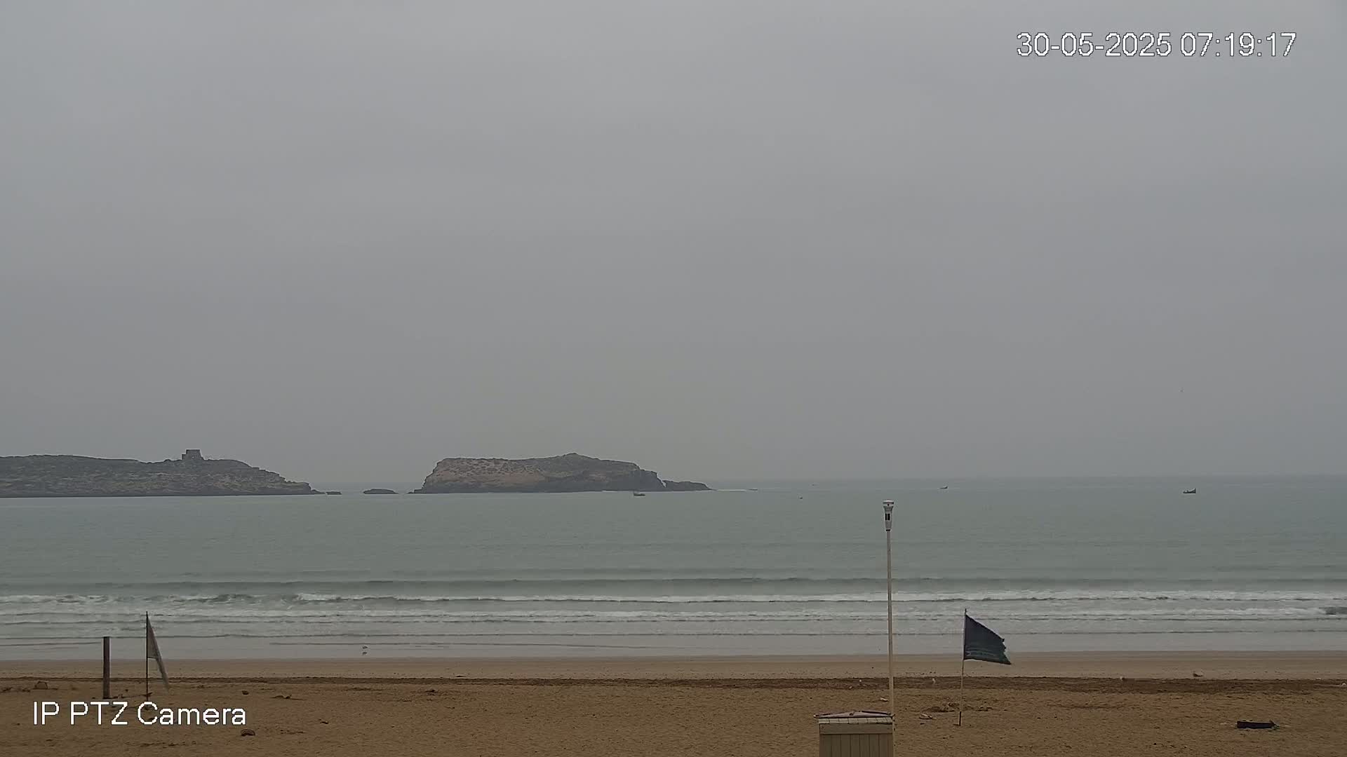 A sandy beach with gentle waves rolls toward two small islands under an overcast sky.