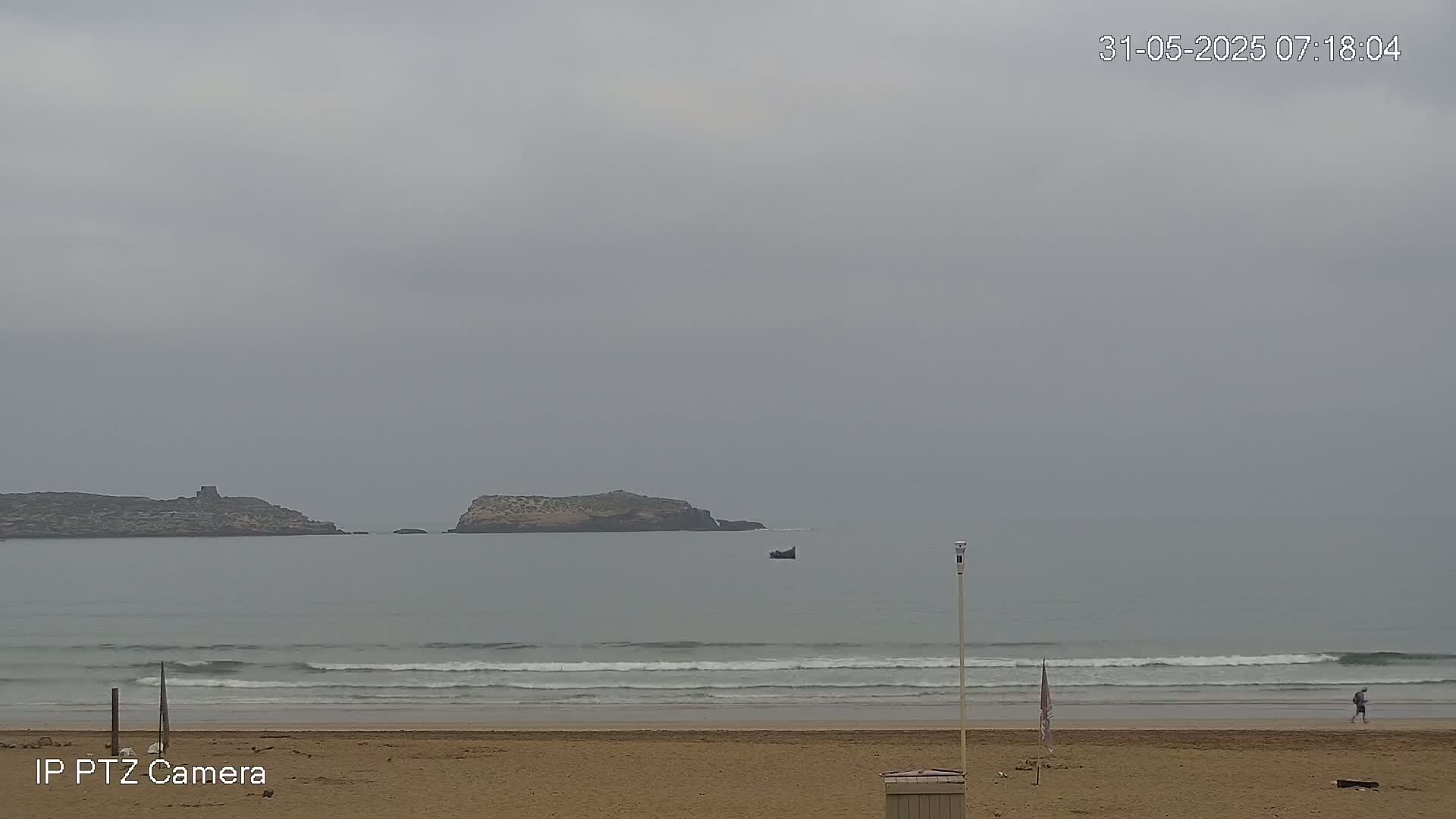 A sandy beach with gentle waves under an overcast sky, featuring two small islands in the distance and a lone person walking along the shore.