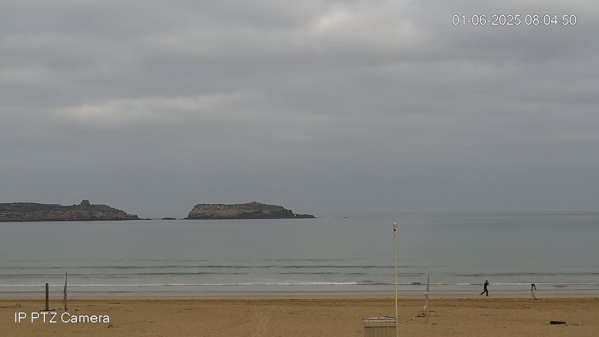 A sandy beach with small waves under an overcast sky features two small islands in the distance and a few people walking on the shore.