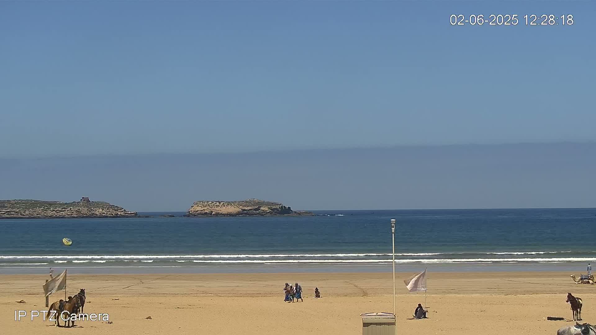 A sandy beach with people and horses, under a clear blue sky, overlooks a calm ocean with small islands in the distance.
