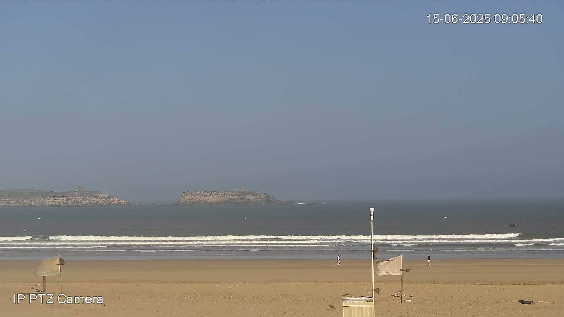 A sandy beach with a few people walking on it and gentle waves rolling in on a clear day.