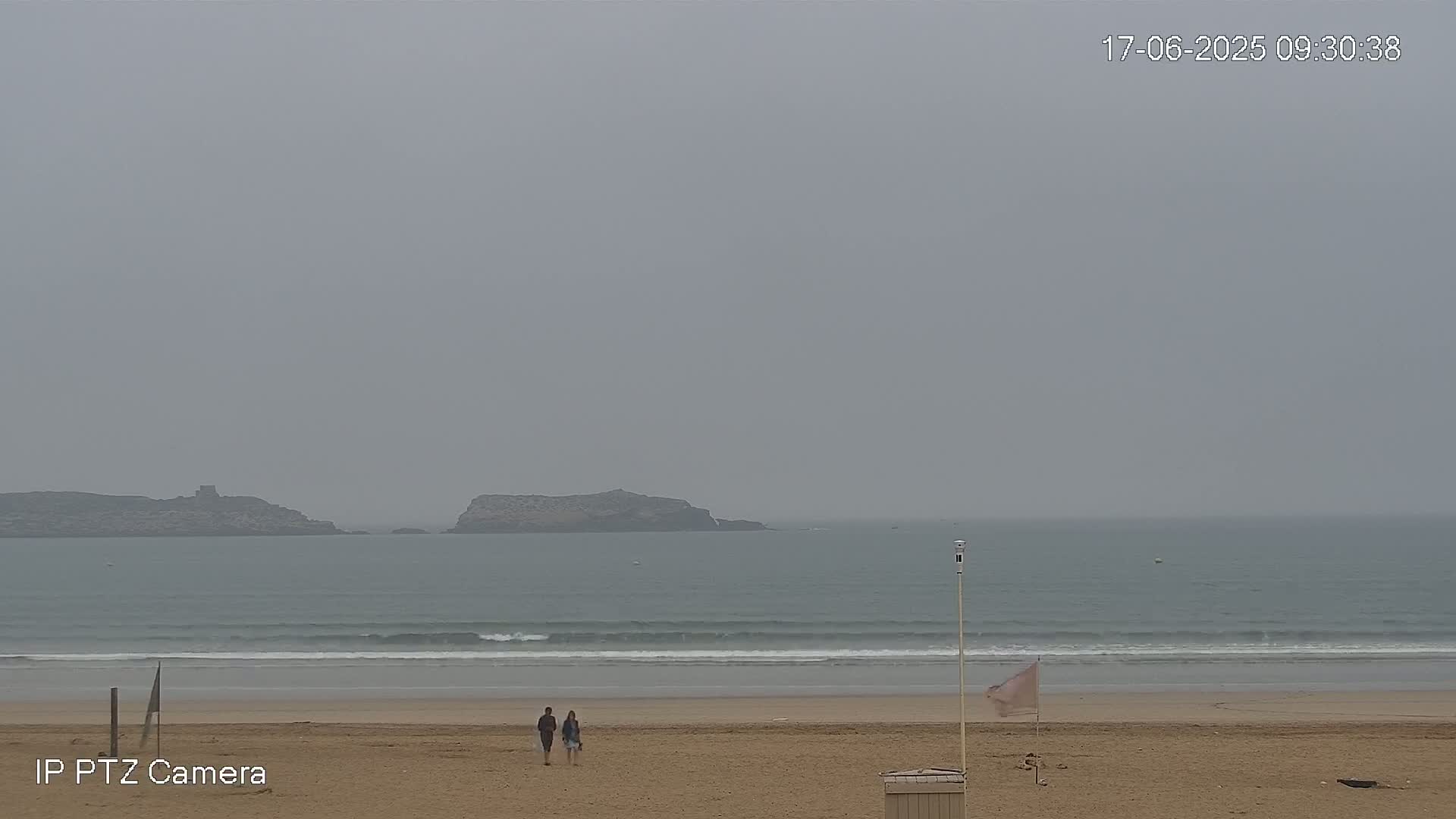 Two people walk on a sandy beach under an overcast sky, toward a calm ocean with small waves and distant islands.