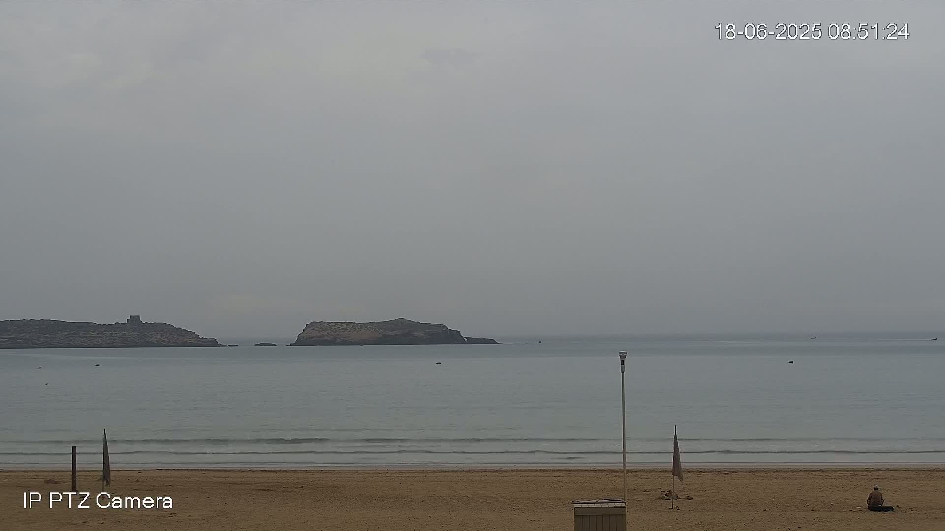 A person sits on a sandy beach overlooking a calm ocean with two small islands visible in the distance under an overcast sky.