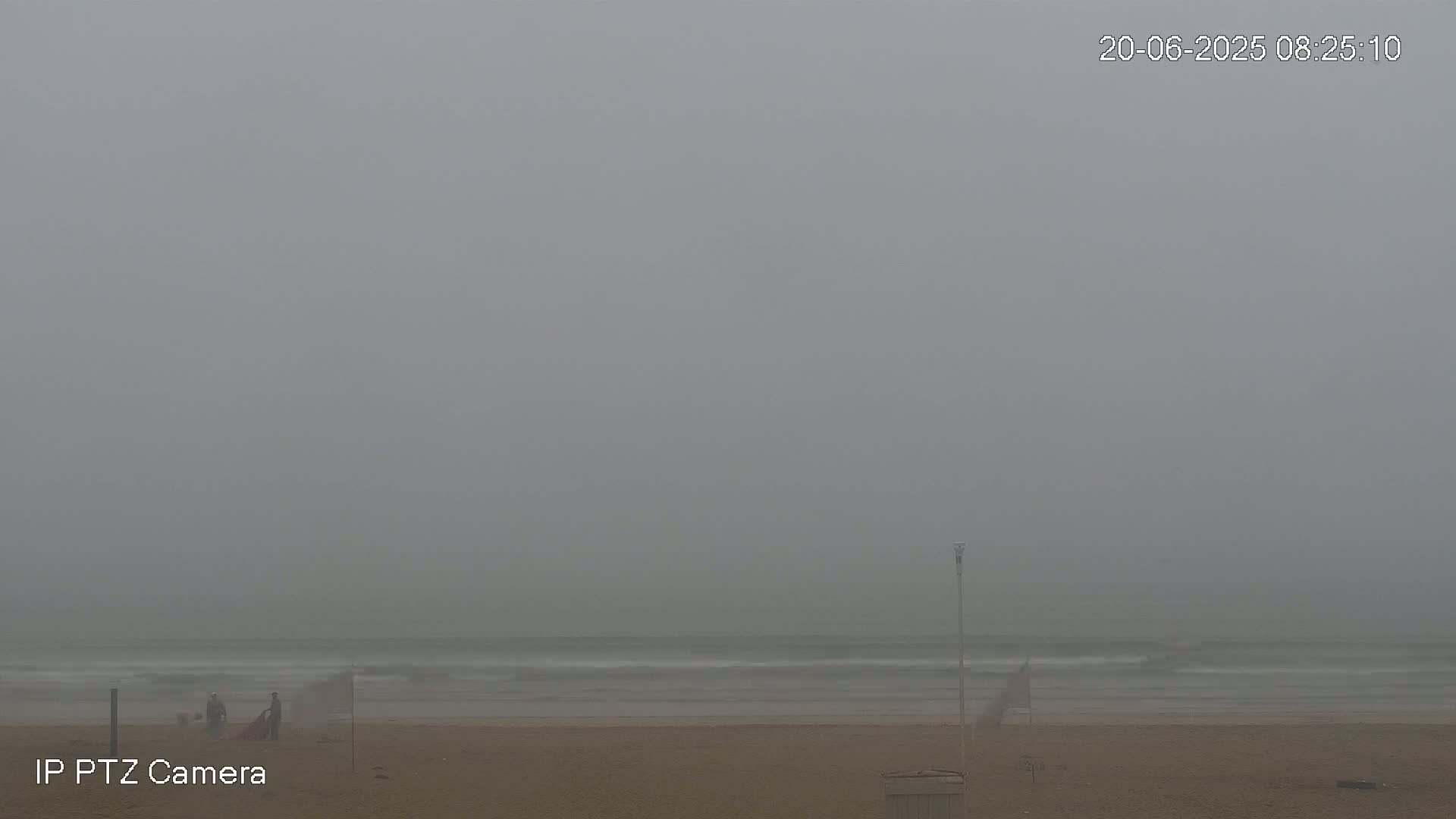 A foggy beach scene shows a few people near the water's edge on a sandy shore.