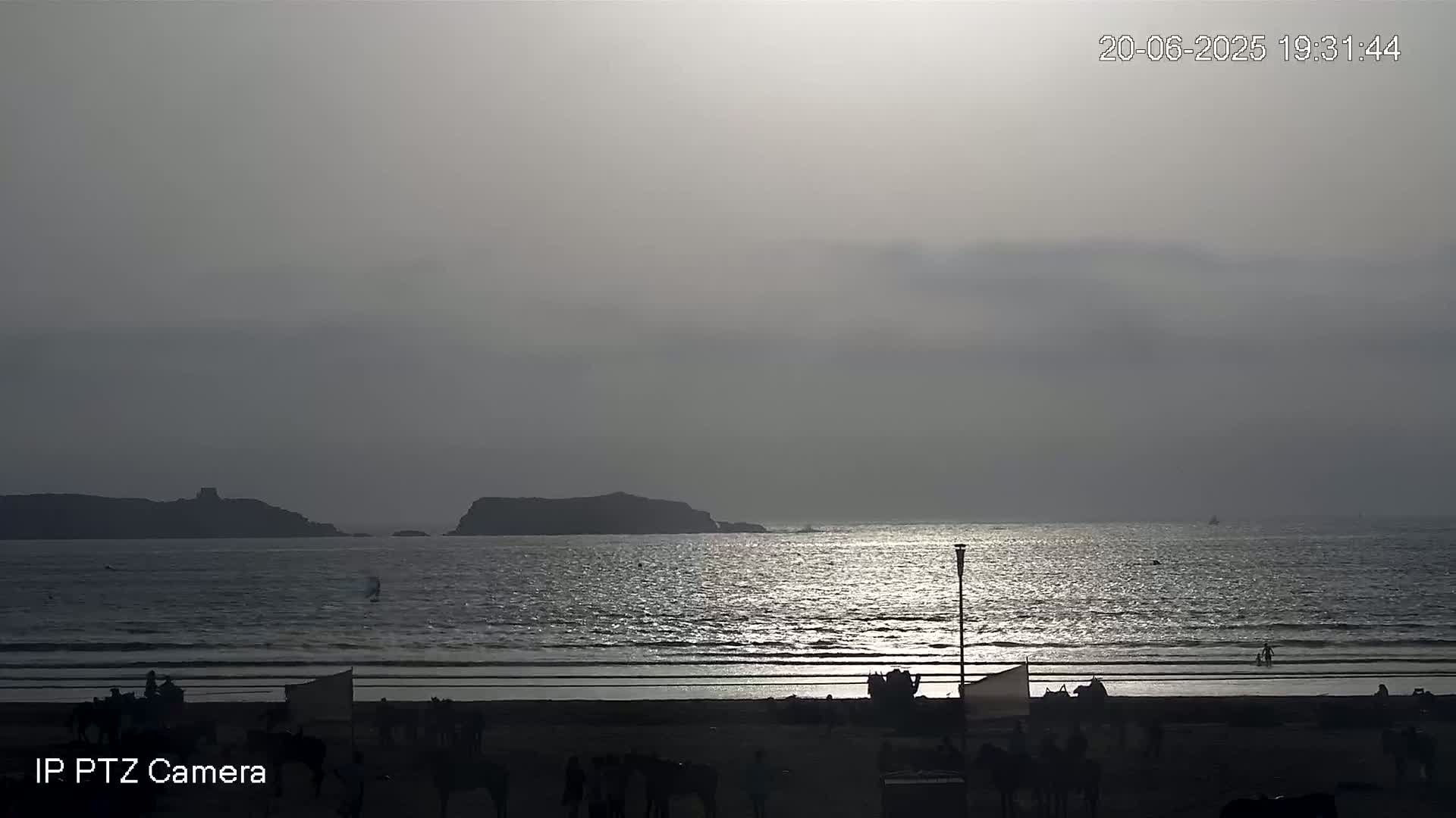 A beach scene at sunset shows a calm sea reflecting the light, with dark silhouettes of landmasses in the distance and people gathered on the shore.