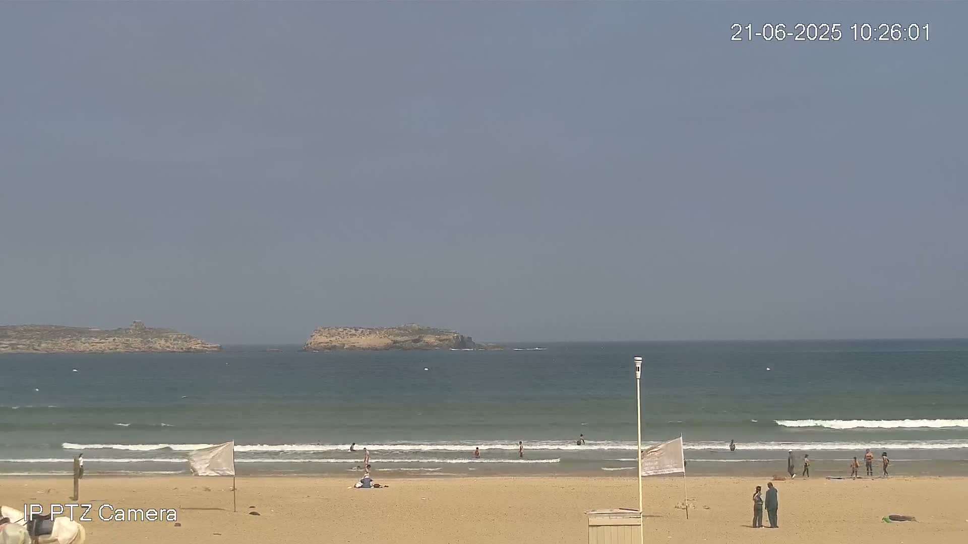 A sandy beach with a few people swimming and relaxing, under a clear sky, with small islands visible in the distance.