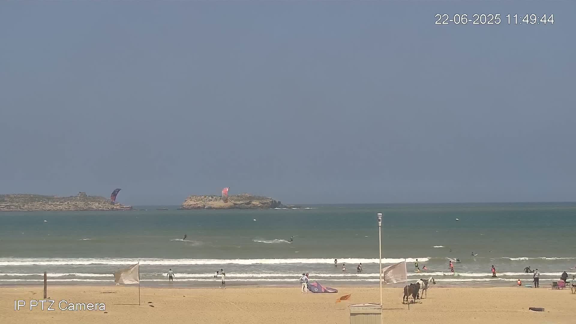 A sandy beach with people swimming and surfing in the ocean under a clear, sunny sky, with two small islands visible in the distance.