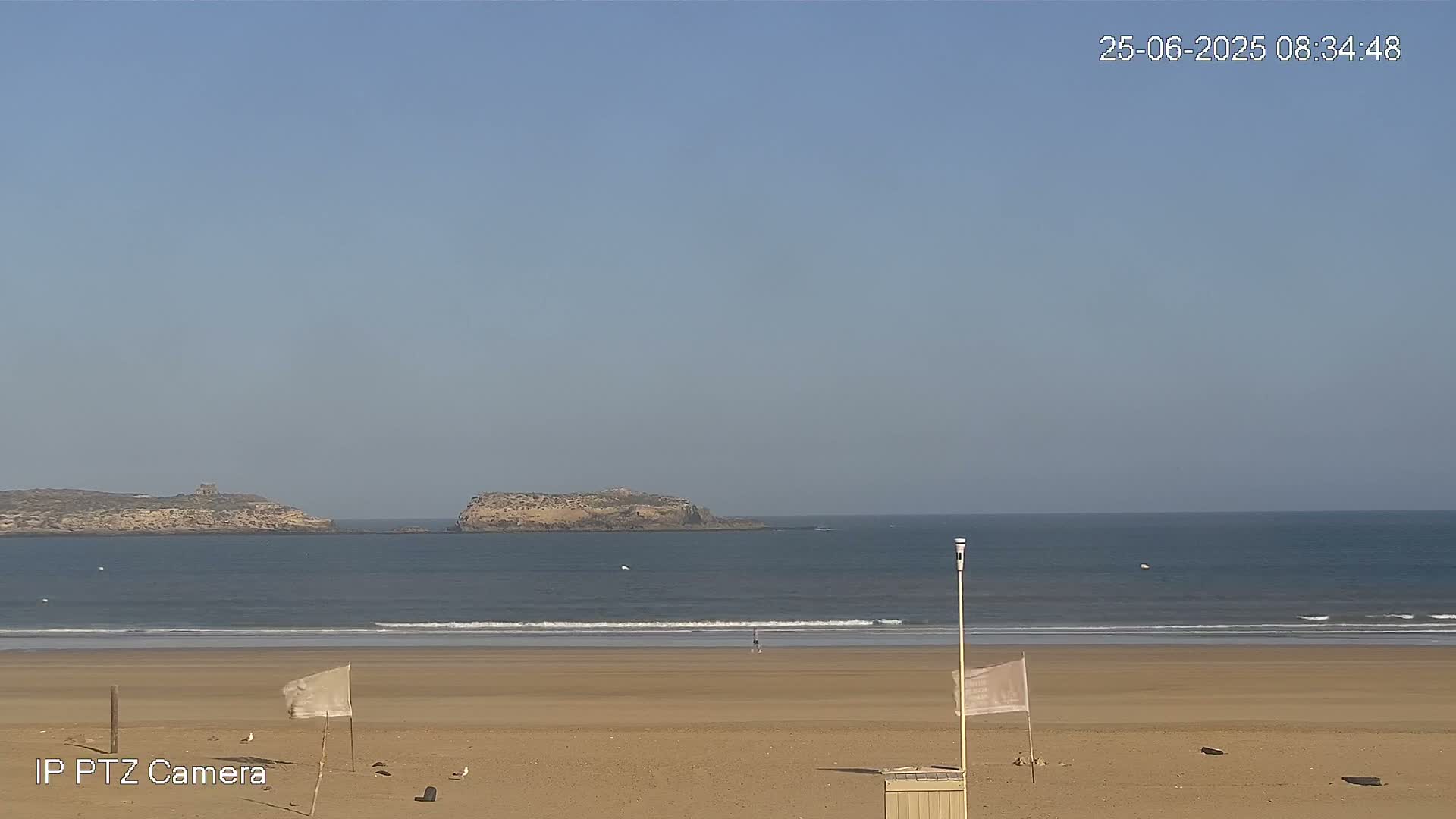 A sandy beach stretches towards a calm ocean with small waves, and two small islands are visible in the distance under a clear sky.