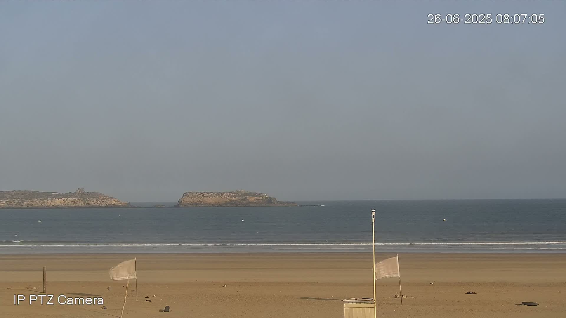 A sandy beach stretches to a calm ocean with two small islands in the distance under a hazy sky.