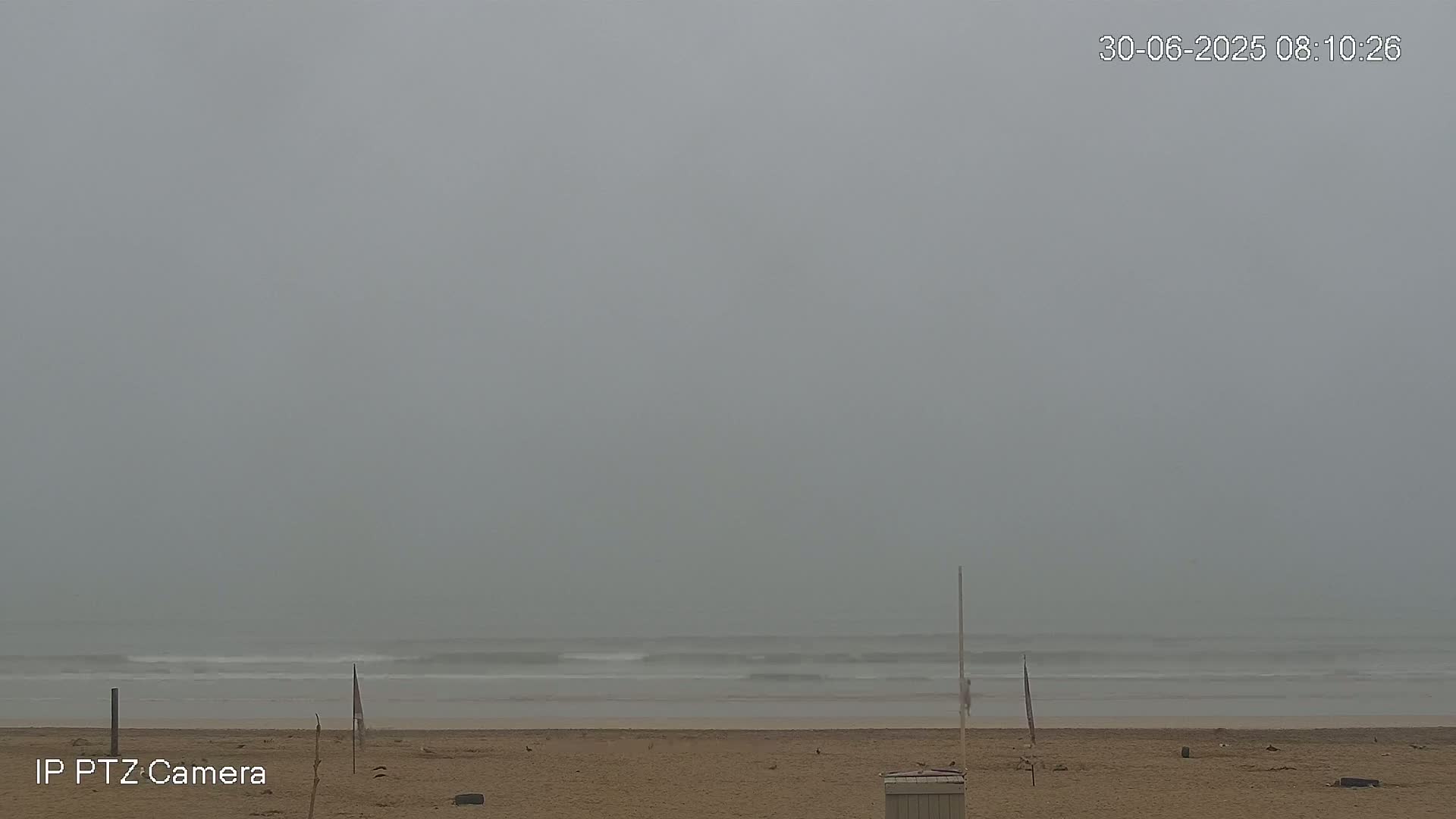 A foggy beach scene shows a sandy shore with small waves and a few flags in the distance.
