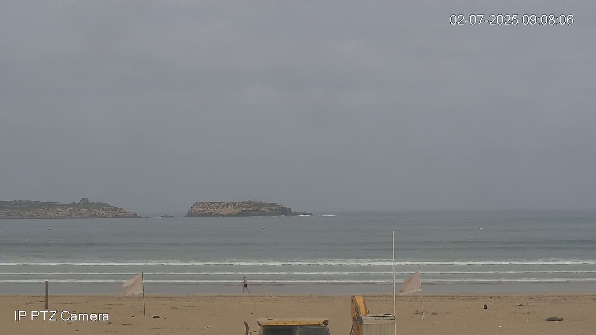 A person walks on a sandy beach under an overcast sky, with small waves rolling into shore near two small islands in the distance.