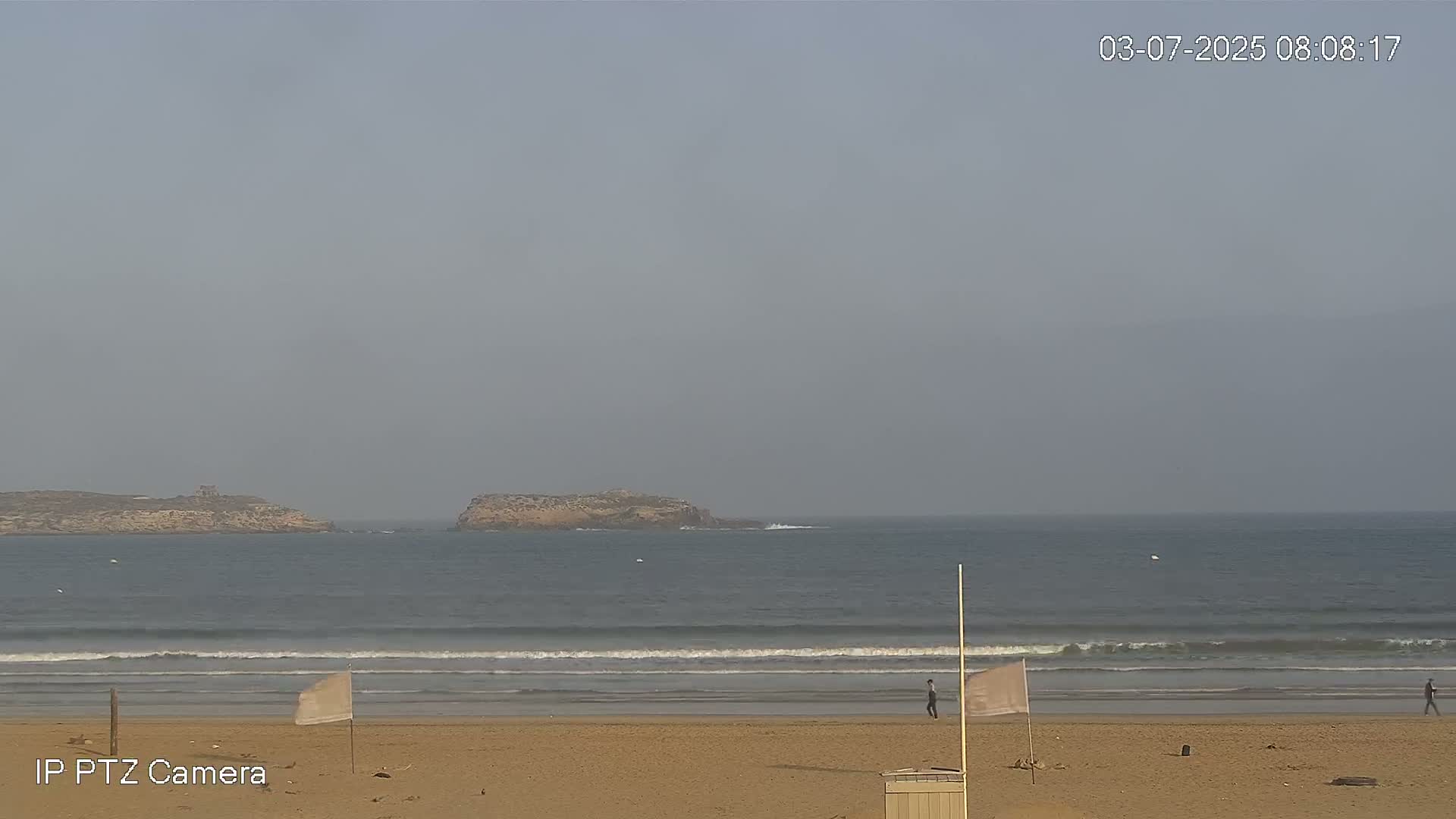 A sandy beach with gentle waves and two small islands visible in the hazy distance.