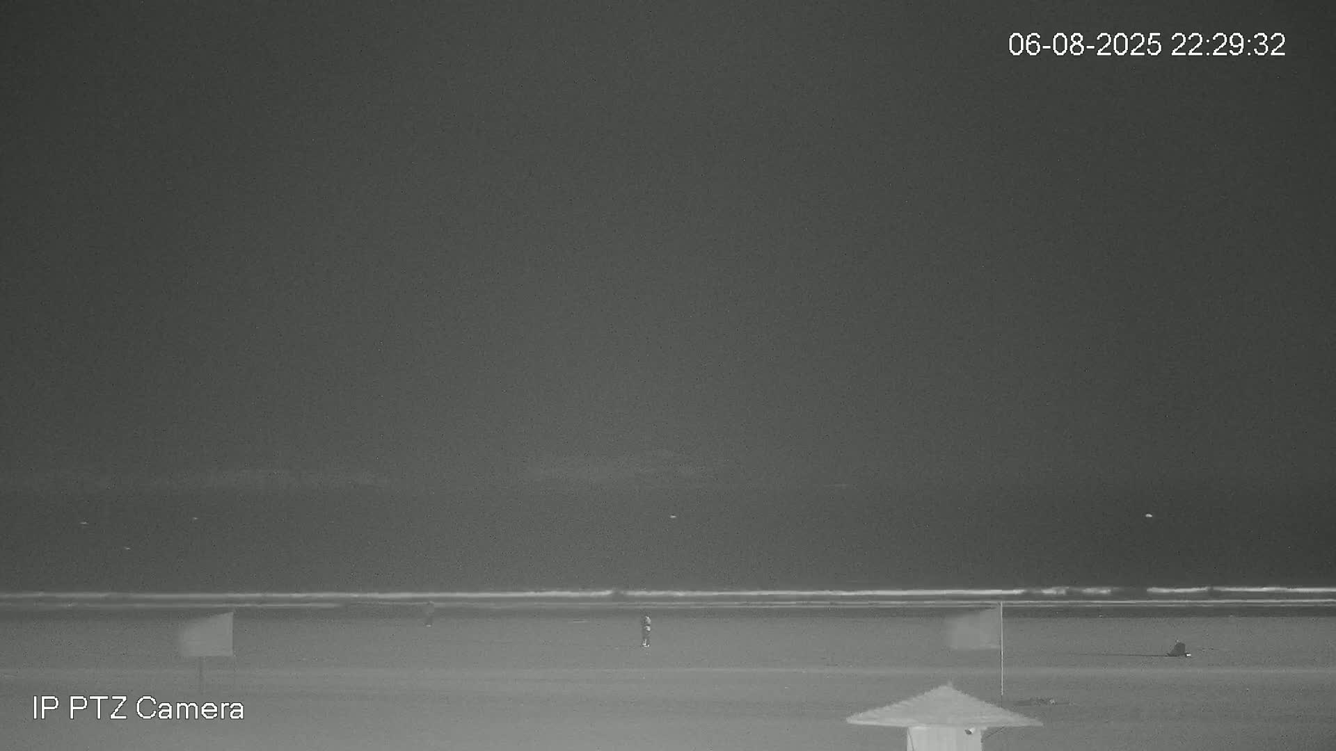 Under a dark night sky, a mostly empty beach is shown with a few people and beach markers, and a small portion of an umbrella is visible in the bottom right corner.