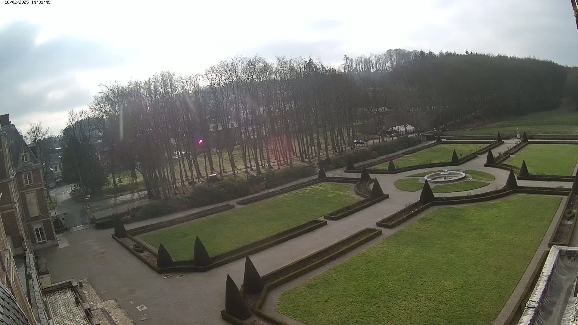 An aerial view shows an ornate, geometrically designed garden with a central fountain, surrounded by a large expanse of green lawn and a backdrop of bare winter trees under a mostly cloudy sky.