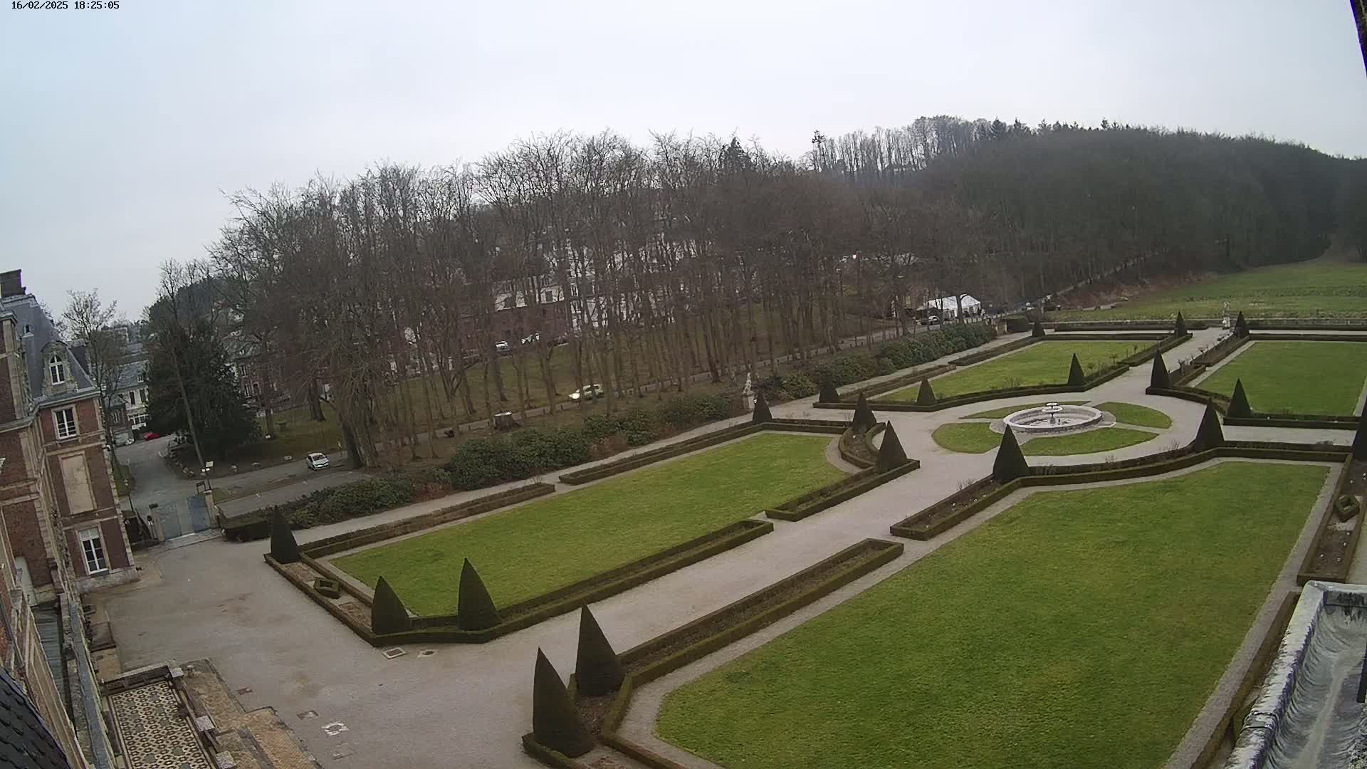 An elevated view shows a formal garden with geometrically arranged lawns, hedges, and a central fountain, situated next to a chateau on an overcast day.