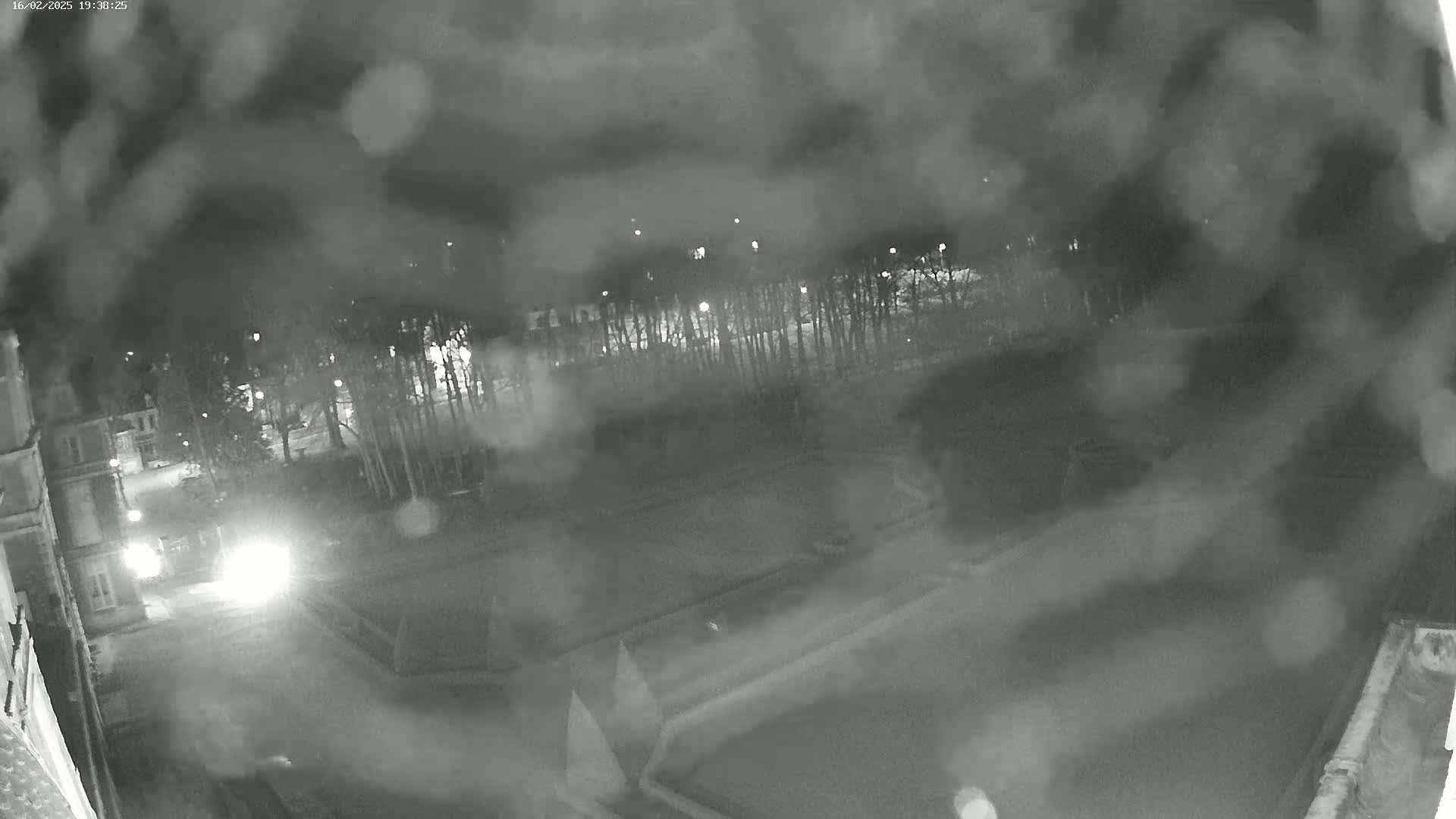 A nighttime, rain-streaked view shows a dark roadway leading to a building and a tree-lined area beyond, with a dark rectangular feature in the foreground.