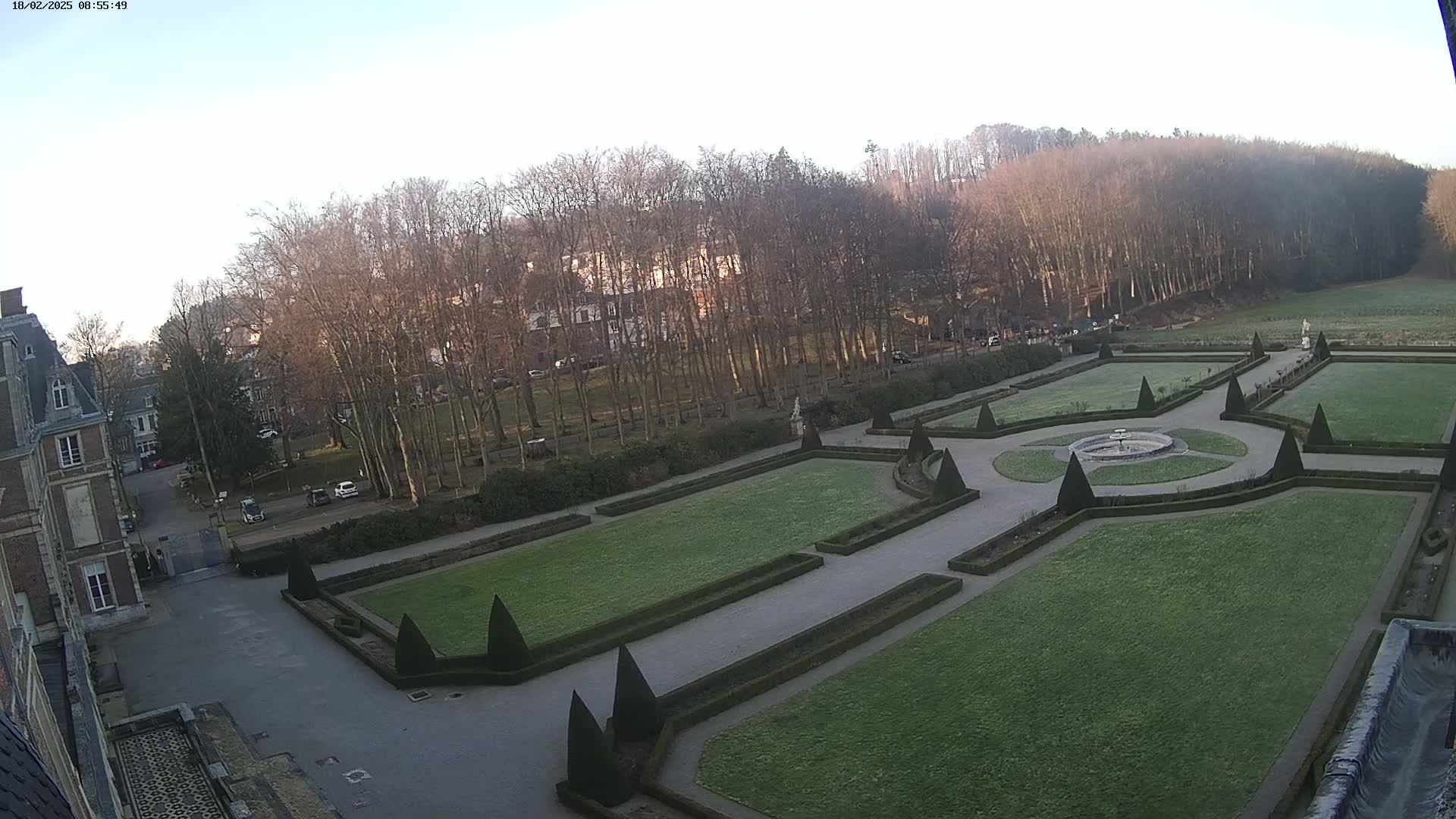 A formal garden with manicured lawns and hedges is situated beside a large building under a clear, bright sky.
