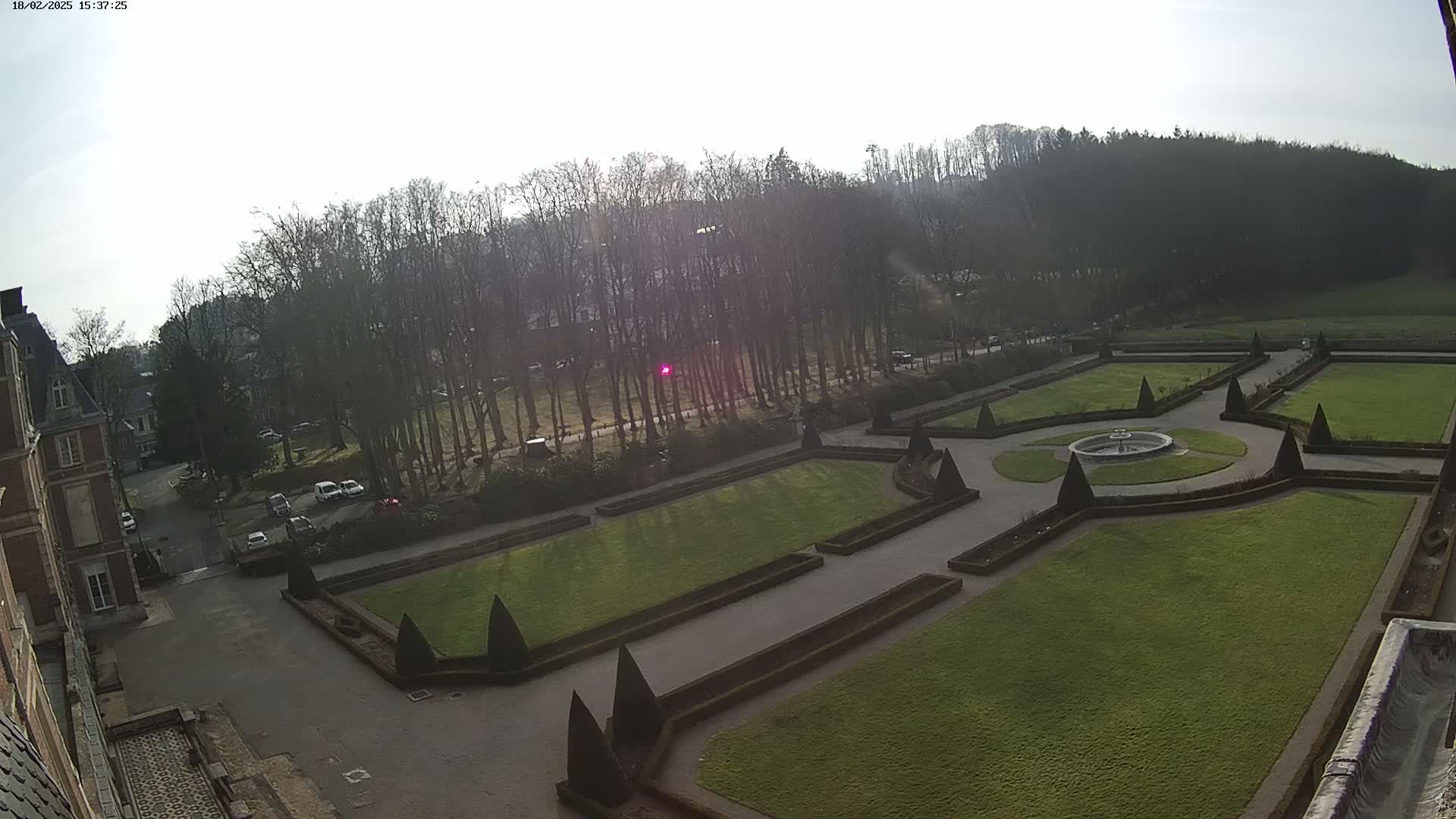 A formal garden with a central fountain is visible from an elevated vantage point on a partly sunny day, next to a building and a line of bare trees.