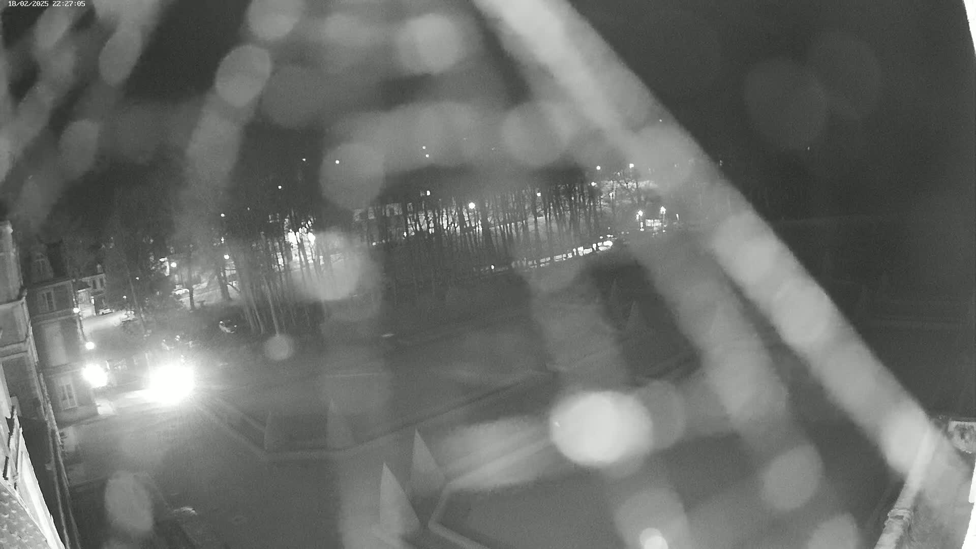 A blurry nighttime view through rain-covered glass shows a dark street with some lights and trees, possibly a parking lot, and buildings in the distance.