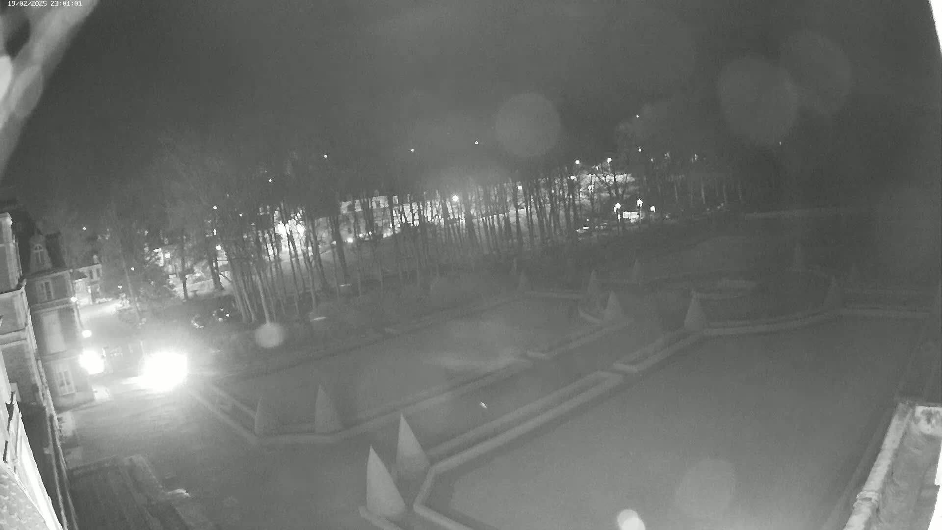 A nighttime, high-angle view shows a formal garden with geometrically designed hedges and pathways, situated near a building and trees under a dark sky.