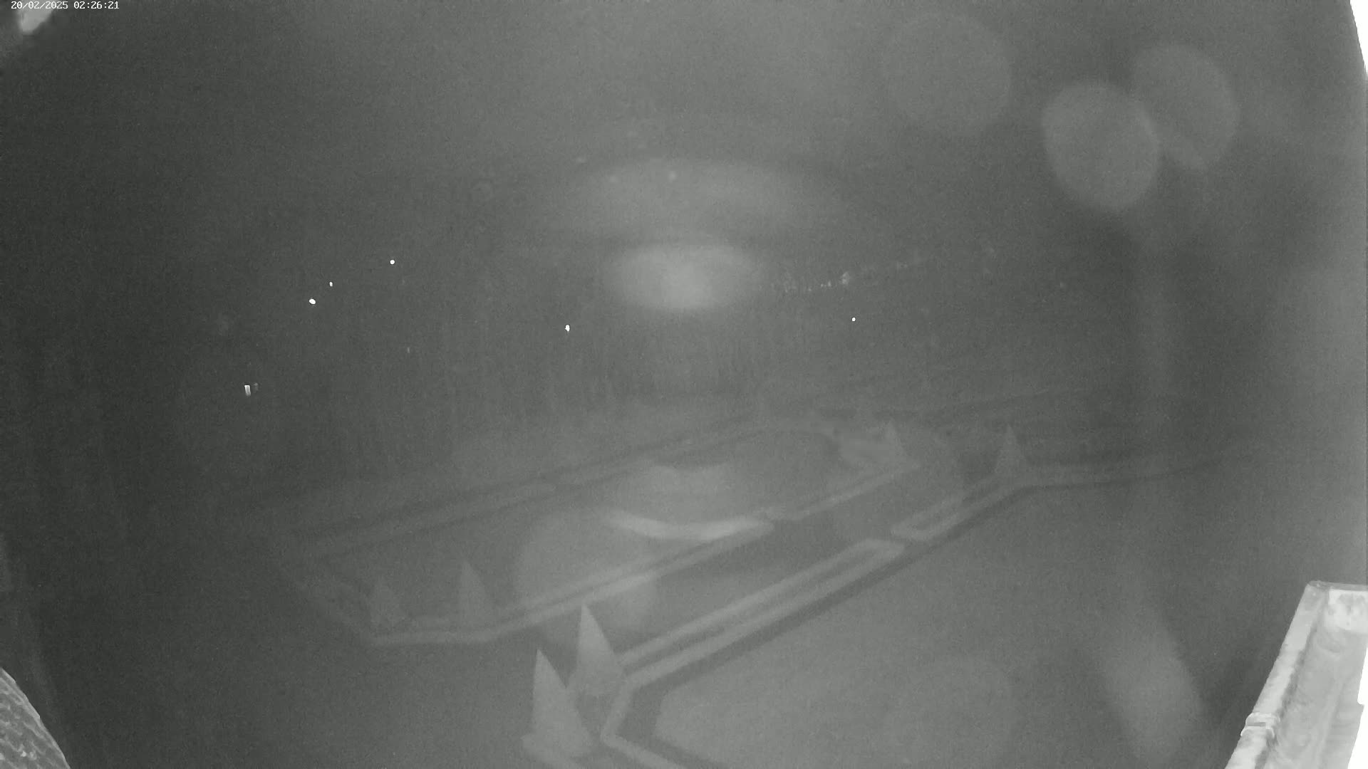 A nighttime, high-angle, low-light view shows a geometric pattern of dark landscaping, possibly a garden or park, with a few faint lights visible in the distance.
