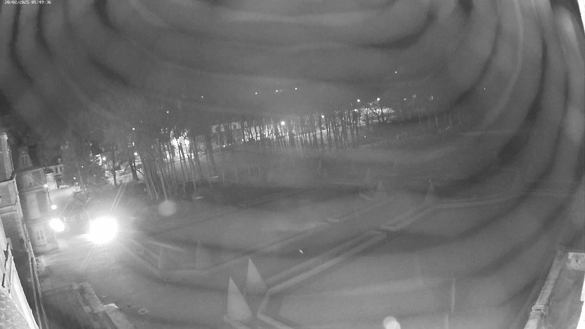 A nighttime, blurry, black-and-white view of a paved area with some kind of landscaping features, trees in the background, and a vehicle's headlights visible in the foreground.