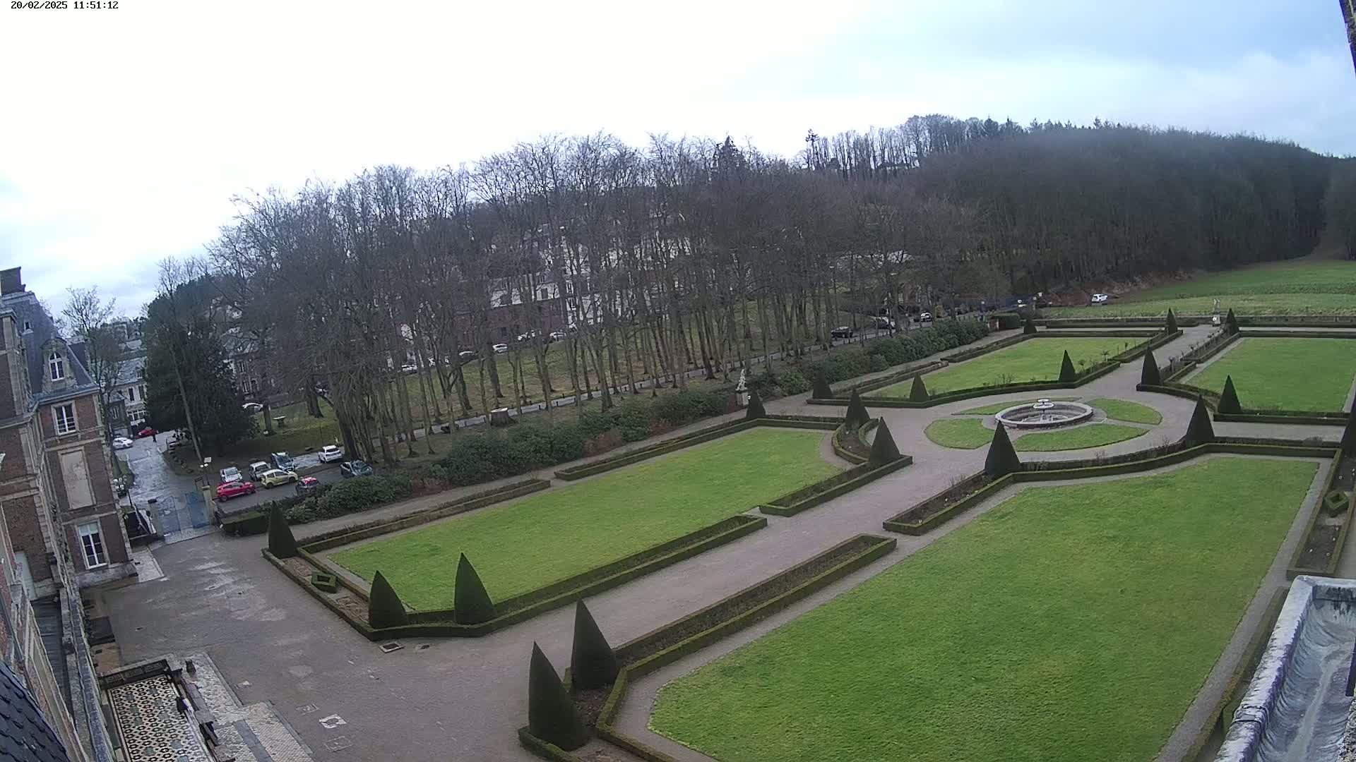 An aerial view shows a formal garden with manicured lawns, hedges, and a central fountain, situated next to a large building and a wooded hillside under a cloudy sky.